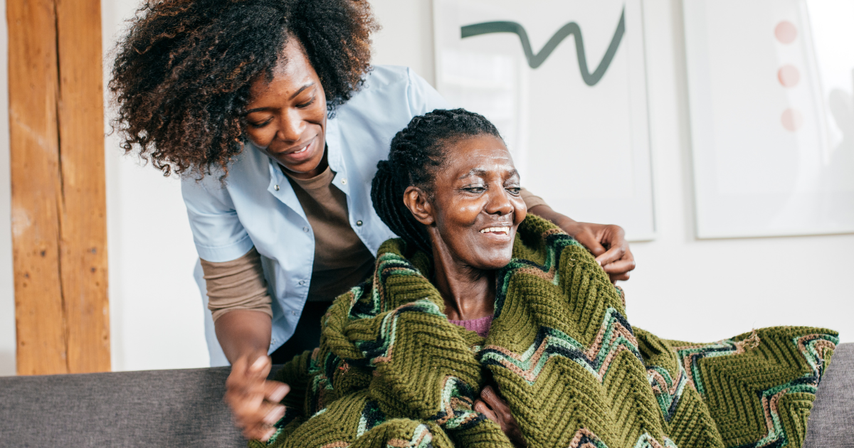 A compassionate caregiver gently comforts a senior woman wrapped in a blanket at home, offering emotional support and companionship.