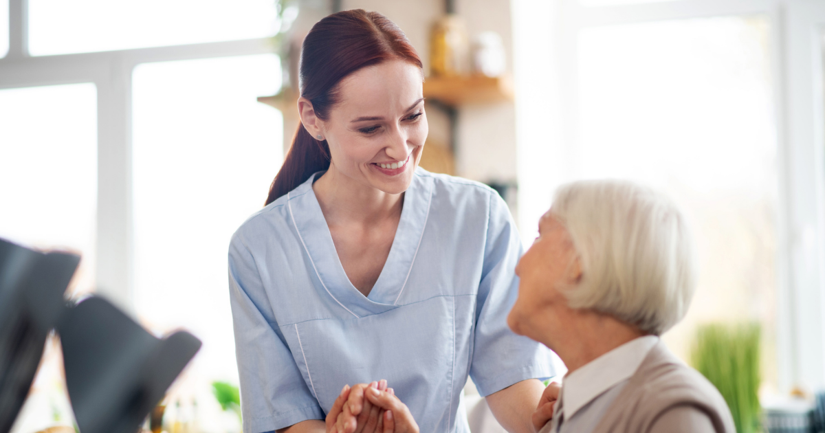A professional caregiver holds the hand of a senior woman during an in-home care visit, offering reassurance and emotional support.