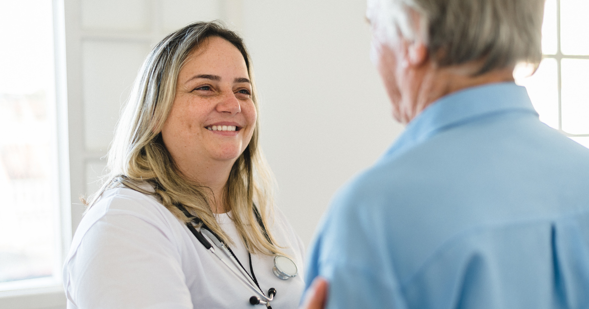 A home health nurse smiles while speaking with a senior man during an in-home visit, offering skilled medical care and support.