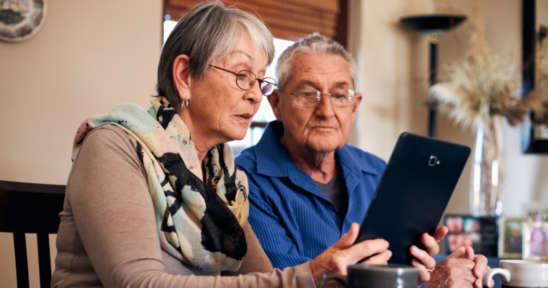 Seniors Using a Tablet Together with In-Home Support An elderly couple sits together at home while reviewing information on a tablet, reflecting technology assistance and supportive senior care.