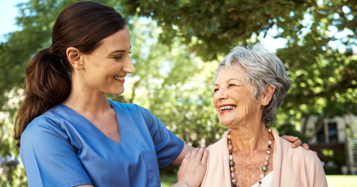 A professional caregiver smiles while offering supportive companionship to a senior woman outdoors, promoting emotional well-being and independence.
