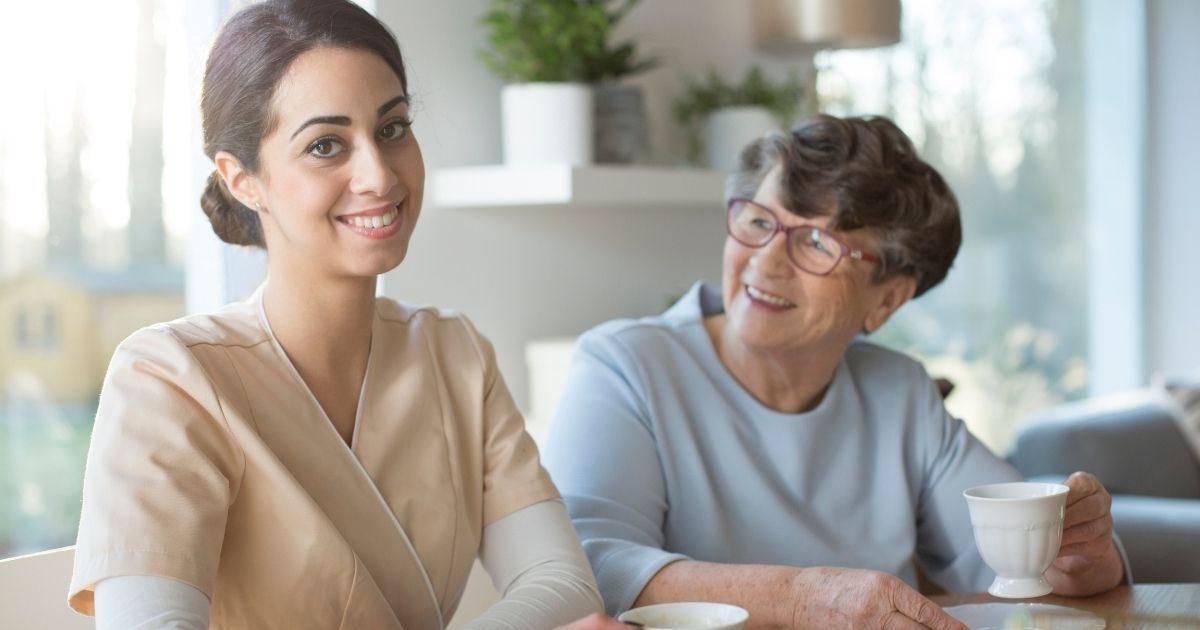 A professional caregiver sits with a senior woman at home, sharing a friendly conversation while enjoying a cup of tea together.