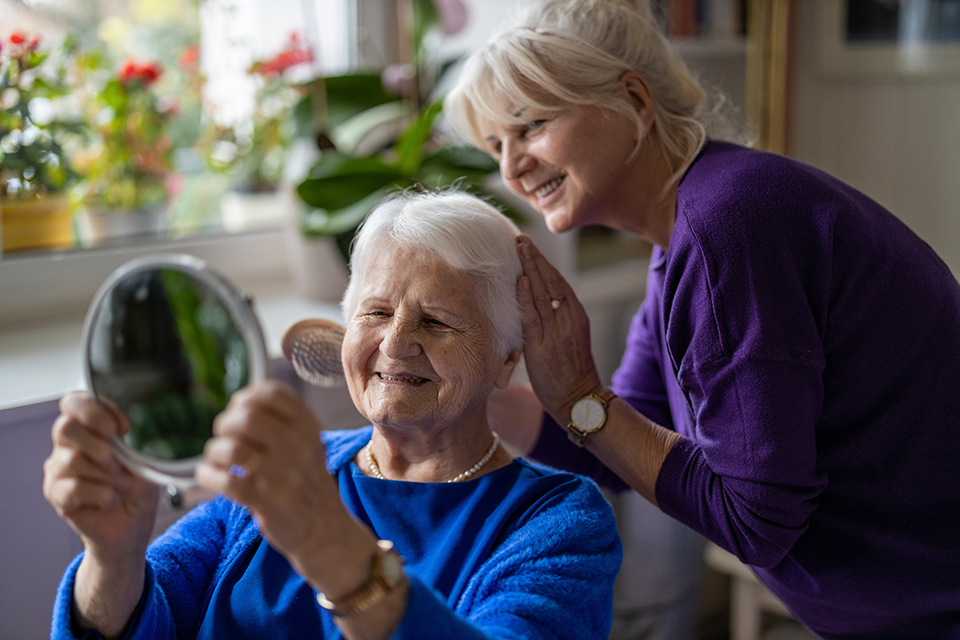 A smiling elderly woman holding a mirror while a caregiver helps adjust her hair, creating a joyful and supportive moment.