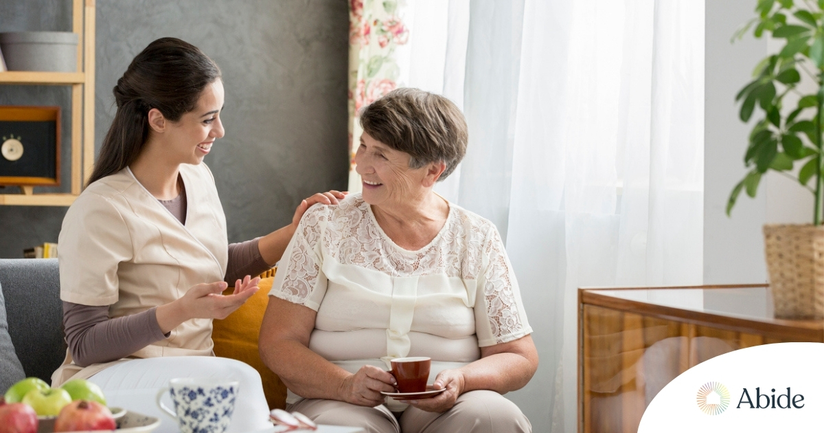 Smiling caregiver with her hand on an older woman’s shoulder as they share tea on the couch, showing home care vs assisted living